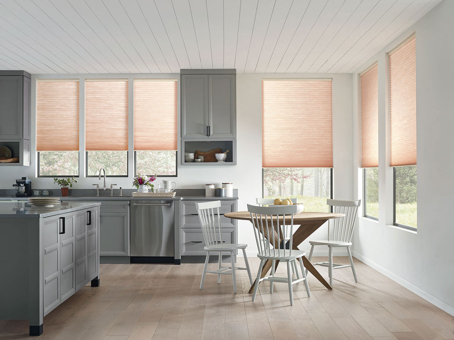 A modern kitchen with grey cabinets, stainless steel appliances, a round wooden table with four white chairs, and large windows featuring Applause® Honeycomb Shades with PowerView® Motorization.