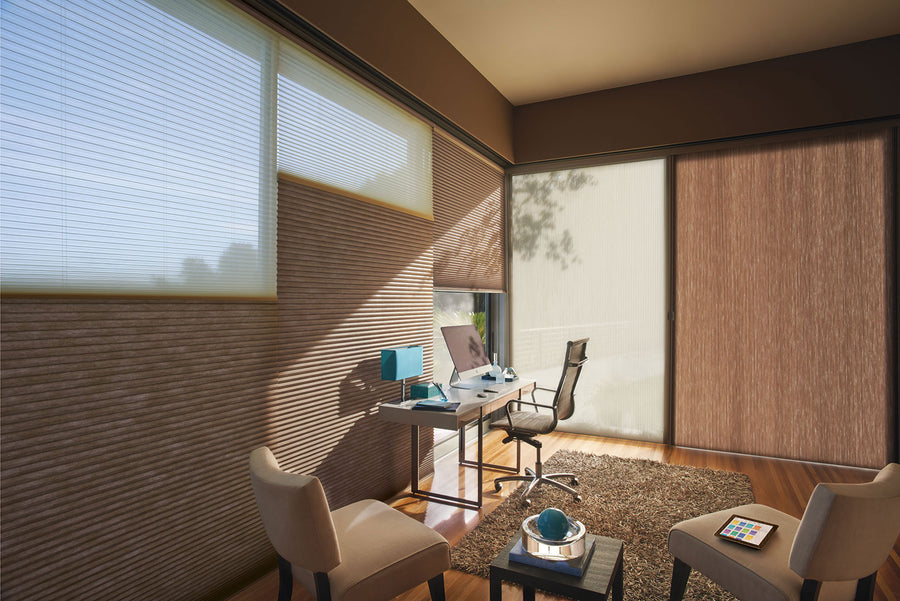 Modern home office with large windows featuring Applause® Honeycomb Shades with PowerView® Motorization, a sleek desk with computer, office chair, two beige chairs, rug, and decorative accents on hardwood floors in natural sunlight.