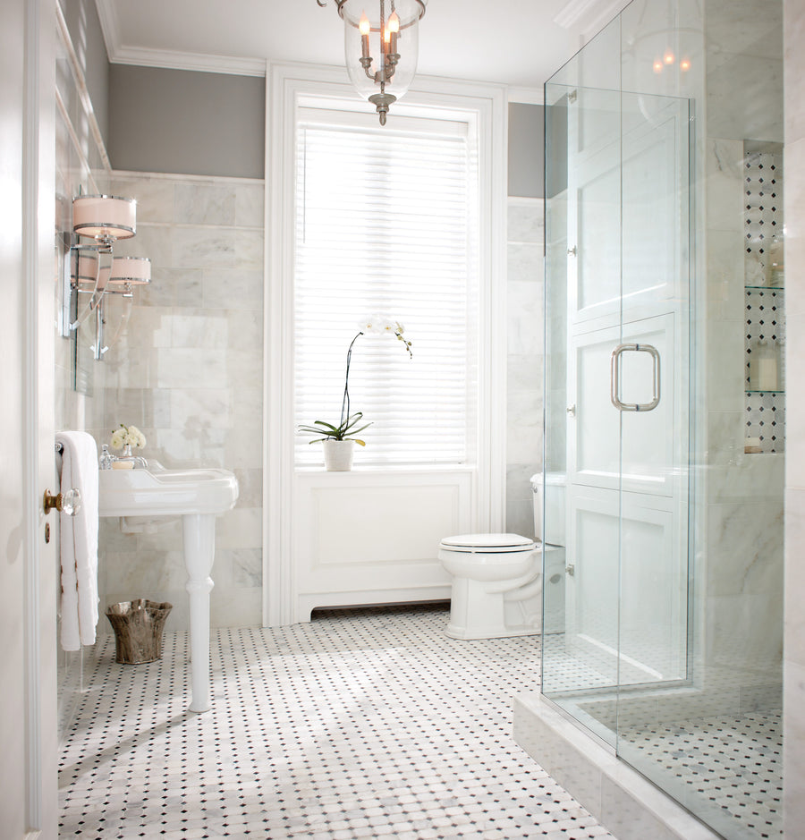 Bright, elegant bathroom with a glass shower, pedestal sink, toilet, Arabescata Carrara Select 6x12 Marble walls, black-and-white tile floor, window with blinds, and a potted orchid on the windowsill.