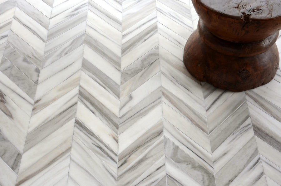 Close-up of Talya Bosphorus 13 7/16 x 13 7/16 Waterjet Natural Stone in a white and gray zigzag pattern; a wooden stool with a round base appears in the upper right, emphasizing the floor’s modern geometric appeal.