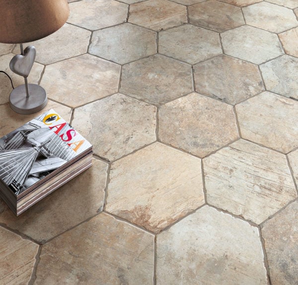 A close-up of a floor featuring Chicago 9x11 Brick Hex Tile, showcasing large, rustic hexagonal porcelain tiles in shades of beige and brown. In the corner are a small table, a decorative object, and a stack of magazines.