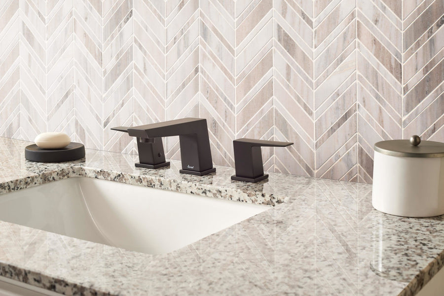 A modern bathroom sink with a granite countertop, matte black faucet, soap dish, and white container set against an elegant herringbone-patterned backsplash made from Chevron Mosaic Natural Stone tiles.