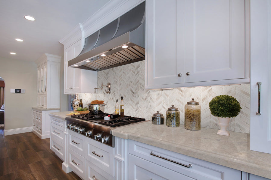 Modern kitchen featuring white cabinets, marble countertops, a gas stove with stainless steel range hood, glass jars of pasta, olive oil bottles, a potted plant, and Chevron Mosaic Natural Stone as backsplash under warm lighting.