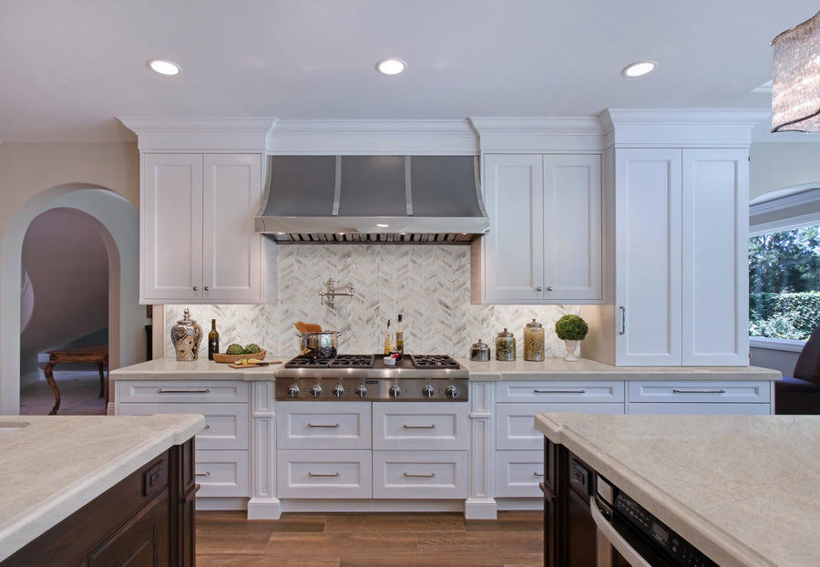 Modern kitchen with white cabinets, stainless steel stove and hood, Chevron Mosaic Natural Stone backsplash tiles, decorative jars on the counters, wooden floor, and bright natural light from a right-side window.