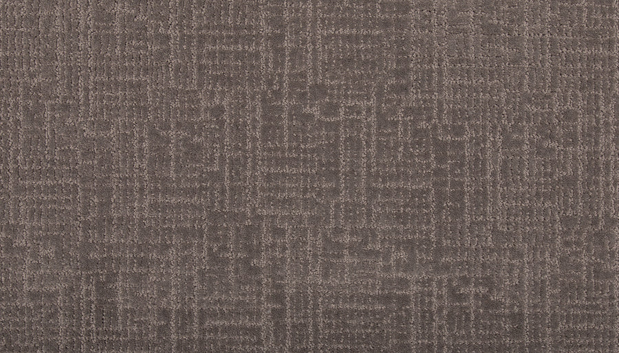 A close-up of Delicate Features Family Friendly Carpet shows its brown color, subtle geometric pattern of intersecting lines and rectangles, soft plush texture, and stain-resistant qualities.