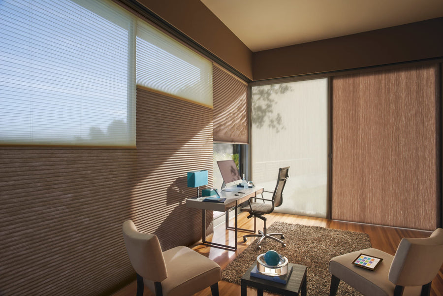 A modern home office with large windows fitted with Commercial Applause® Honeycomb Window Shades (double-cell, light brown), a sleek desk and computer, two beige chairs, a gray office chair, minimal decor, and sunlight gently filtering in.