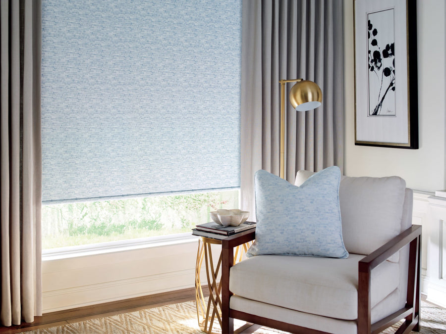 A cozy reading nook with a light armchair, patterned blue pillow, small side table, modern floor lamp, gray curtains, Commercial Designer Roller Shades in blue, and black-and-white floral artwork. Natural light fills the room.