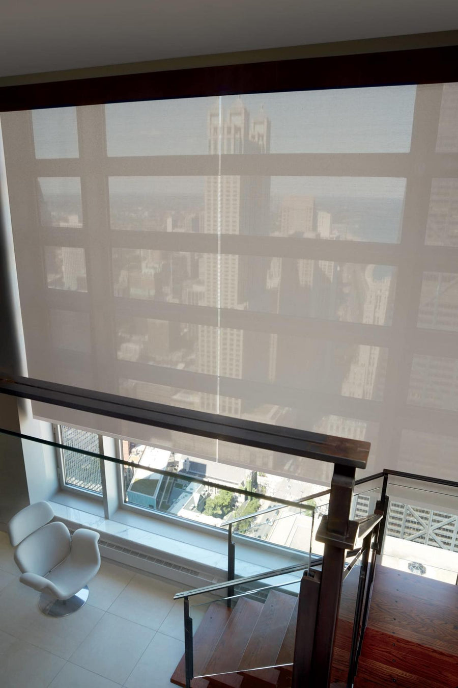 A city view through a large window with Commercial Hunter Douglas Designer Screen Shades offering UV protection. Tall buildings outside, while inside a modern white chair sits beside a glass railing and wooden stairs—perfect for stylish commercial spaces.