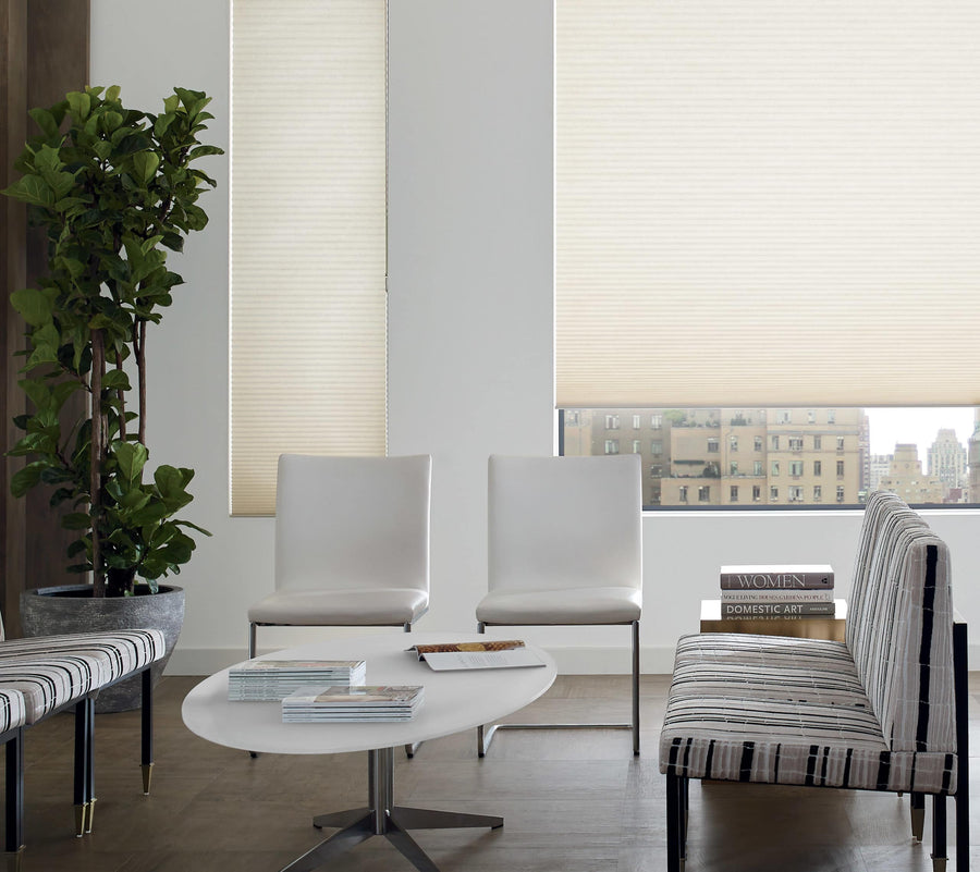 Modern waiting area with white chairs, a striped bench, a round white table with magazines, a potted plant, and large windows fitted with energy-saving Commercial Duette® Honeycomb Shades overlooking city buildings.