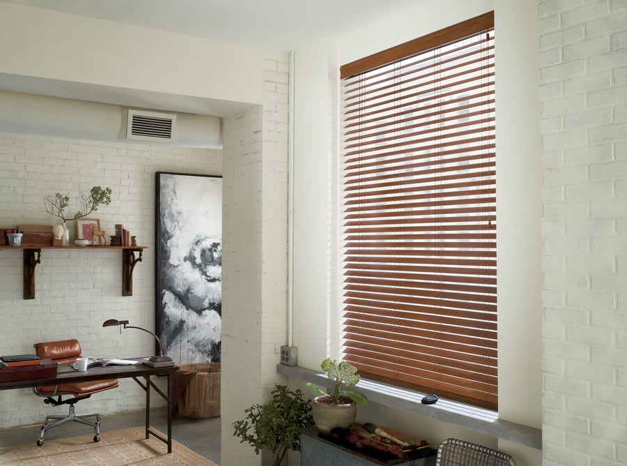 A bright home office with white brick walls, a large window with Commercial Everwood® Alternative Wood Blinds, a brown leather chair at a desk, shelving with decor, potted plants, and a black-and-white abstract painting.