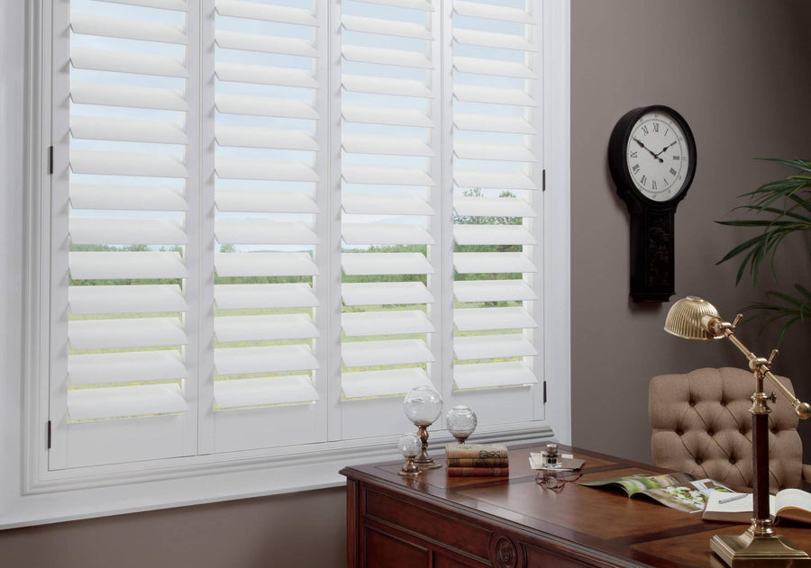 A home office with a large window fitted with Commercial NewStyle® Window Shutters, a wooden desk with papers and a lamp, a tufted chair, wall clock, and leafy indoor plant.