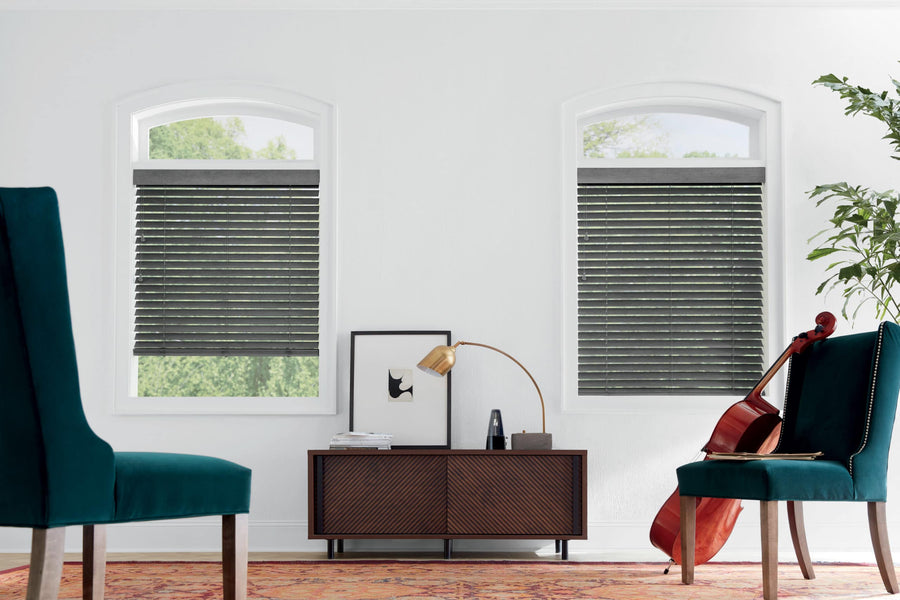 A modern living room features two teal chairs, a low wooden cabinet, a lamp, framed artwork, a potted plant, a cello, and two windows fitted with Commercial Parkland® Wood Blinds that let in natural light.