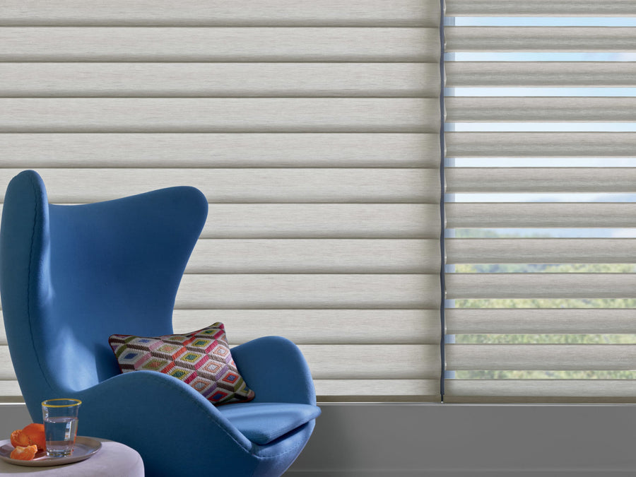 A modern blue armchair with a colorful geometric pillow sits by a small table holding water and fruit. Behind, Commercial Pirouette® Window Shadings offer UV protection while partially revealing the window view.