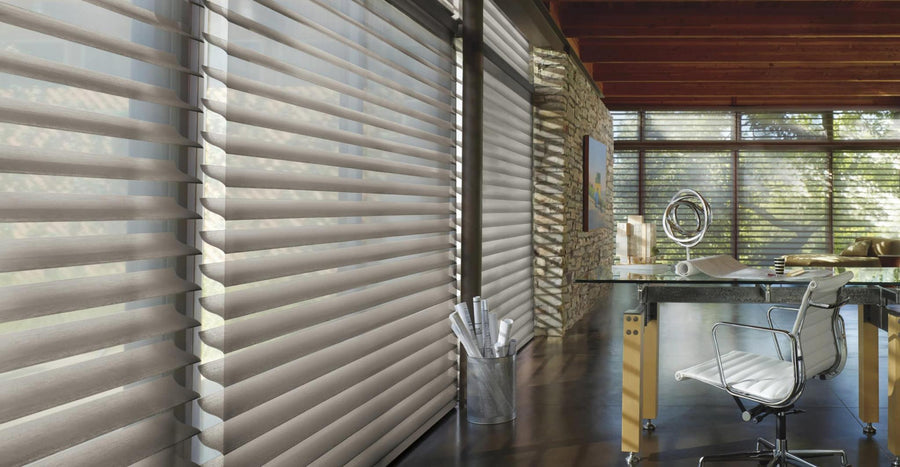 Modern home office with large windows fitted with Commercial Silhouette® Window Shadings, a glass desk paired with a white chair, a metal waste bin holding rolled papers, stone accent wall, and wooden ceiling beams.
