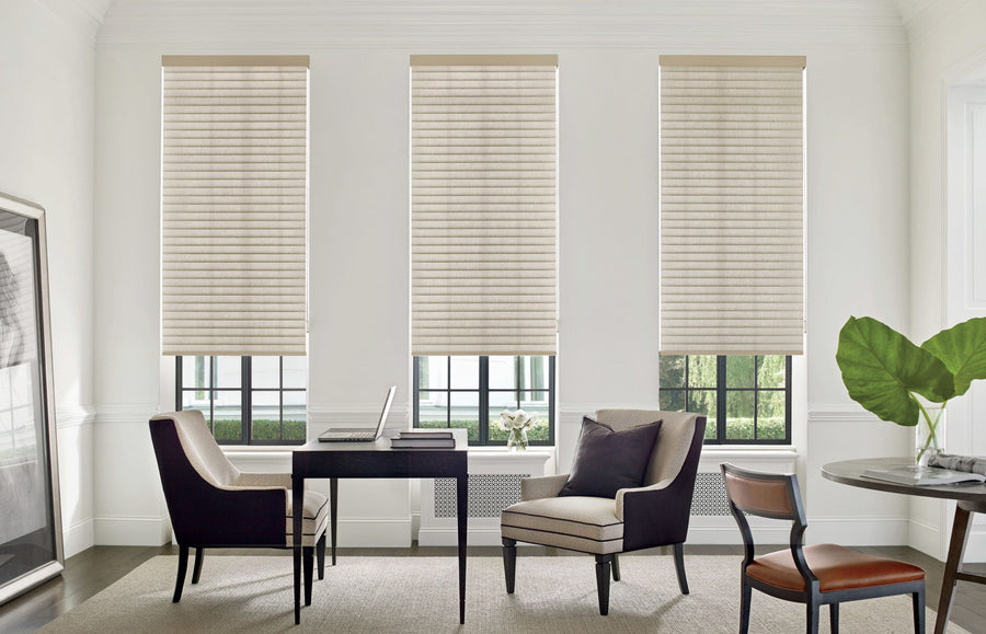 A modern home office with three large windows covered by Commercial Sonnette™ Cellular Roller Shades in beige, a black desk with two chairs, a laptop, a vase of flowers, a framed picture, and large green leaf decor.