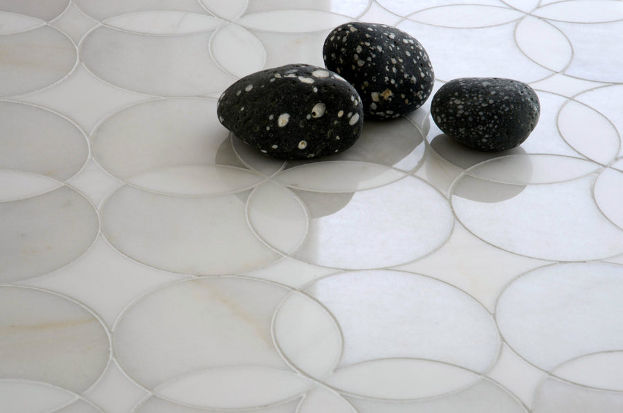 Three black stones with white speckles sit atop large, overlapping Talya Constantine 13 5/8 x 13 5/8 Waterjet Natural Stone tiles, softly lit to highlight their smooth textures and modern geometric patterns reminiscent of marble and stone.