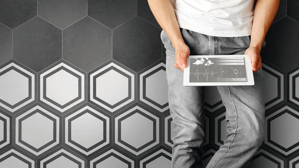 A person in a white shirt and gray jeans stands against a modern wall featuring 8x9 Glazed Hex Tile in varying shades of gray, holding a tablet with charts and graphs.