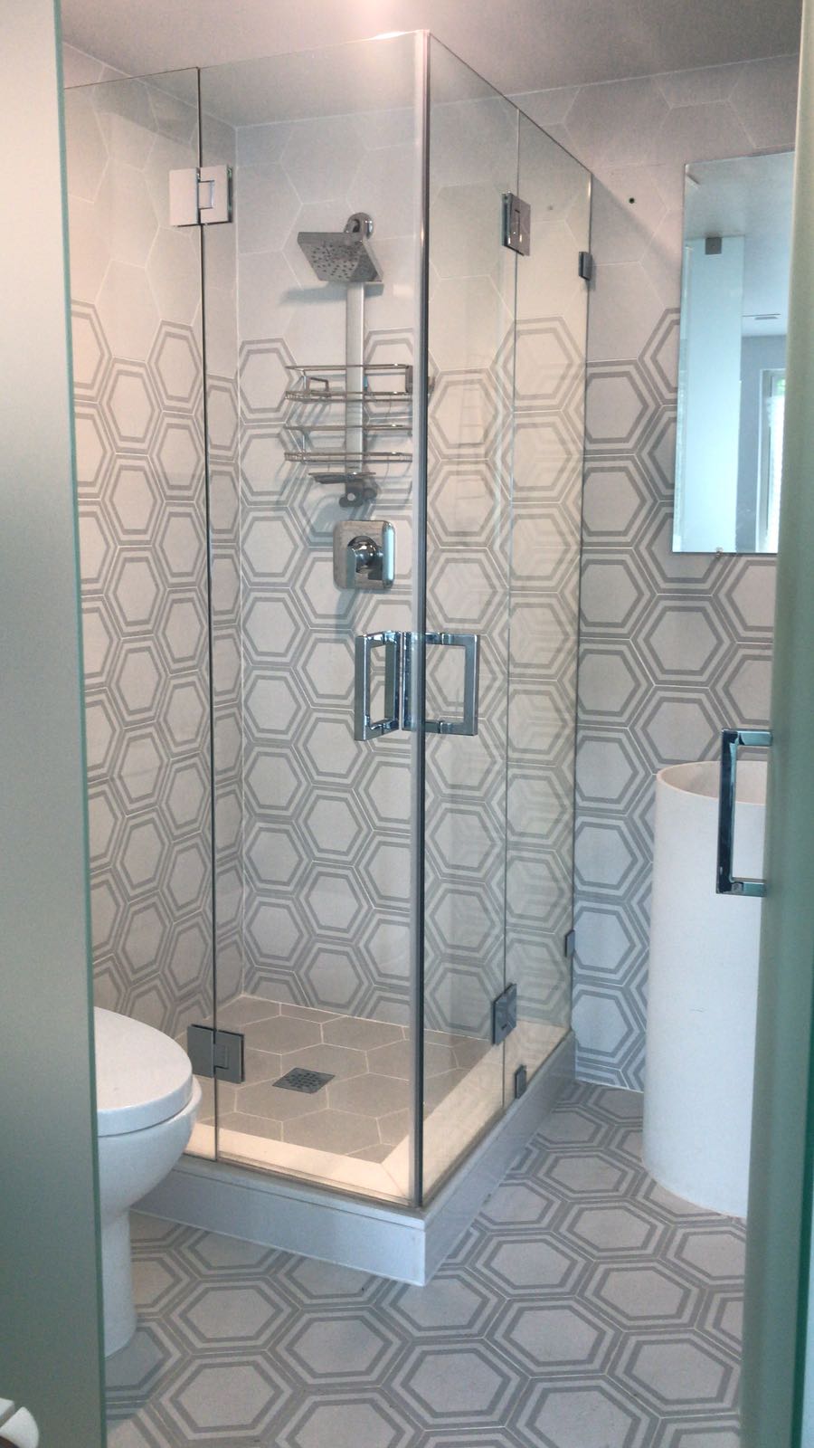 Modern bathroom with a glass-enclosed corner shower featuring 8x9 Glazed Hex Tiles on the walls and floor. A toilet, cylindrical sink, and wall mirror complete the bright, elegantly decorated space.