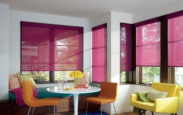 A bright, modern dining nook features large windows with customizable Designer Roller Shades in pink, a round white table with yellow flowers, yellow and orange chairs, colorful cushions, and sunlight streaming in.