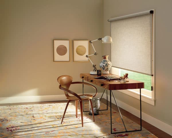 A minimalist home office features a wooden chair, mid-century modern desk, lamp, vase, framed art, patterned rug, and a window dressed with Designer Roller Shades that welcome natural light.