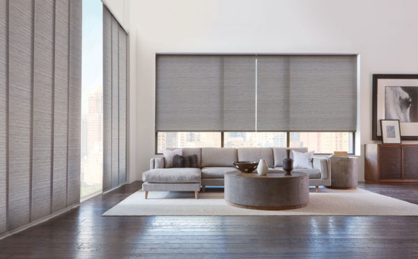 A modern living room features large windows with Designer Roller Shades, a light gray sectional sofa, a round coffee table, and contemporary wall art, offering views of city buildings through the windows.