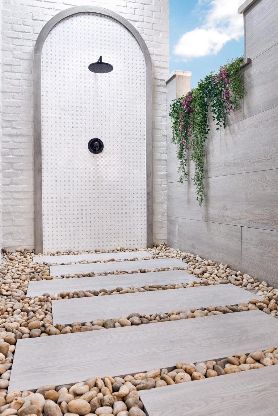 A modern outdoor shower features a white Dolomite Dotty Mosaic Natural Stone wall, black showerhead, stone pathway, and wooden planks with river rocks; green plants hang from a ledge on the right beneath a blue sky.