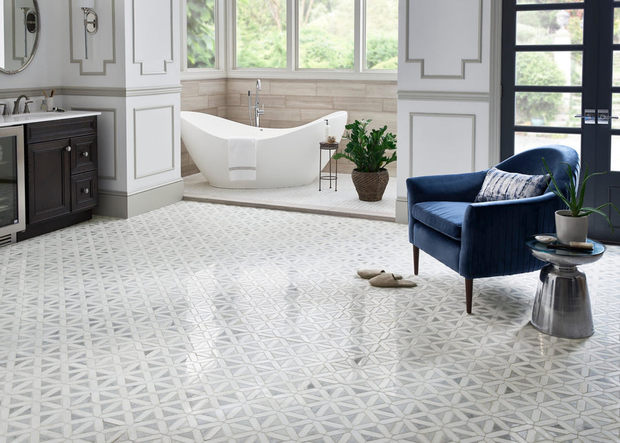 Spacious bathroom with Dolomite Geometrica Mosaic Natural Stone floor, freestanding white tub, dark wood vanity, velvet blue armchair with pillow, small round side table, green plant, slippers, and large windows for natural light.