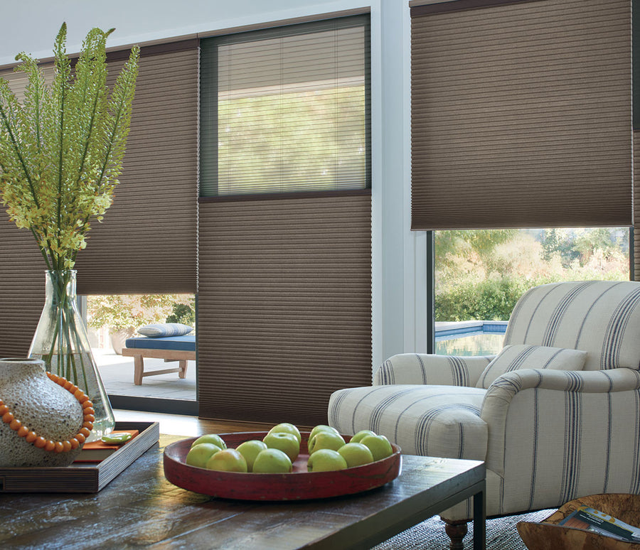 A cozy living room with a striped armchair, a table with green apples, and large windows featuring Duette® Duolite® with PowerView® Motorization window treatments that gently filter sunlight. Outside, trees and a pool are visible.