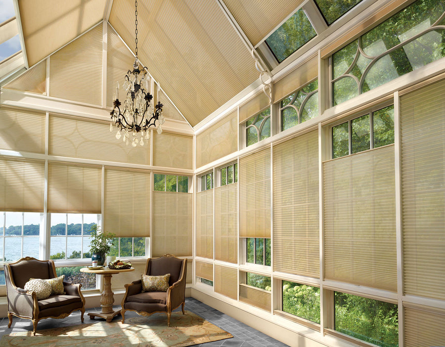 A bright sunroom with large windows adorned with Duette® Honeycomb Shades with PowerView® Motorization, elegant armchairs, a round table set for tea, a patterned rug, and a decorative chandelier. Lush greenery and water can be seen outside.