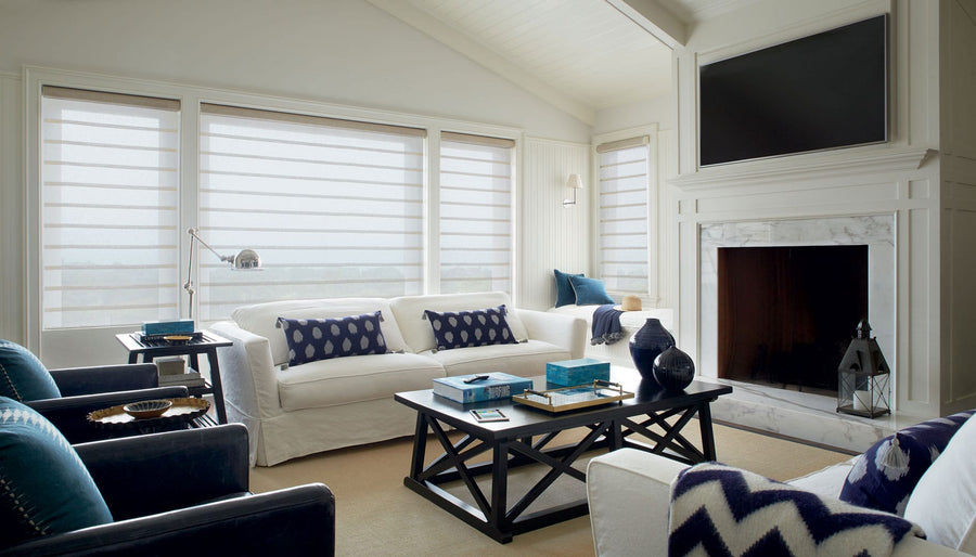 Modern living room with white walls, a large fireplace, and tall windows featuring Duette® Duolite® with PowerView® Motorization. White sofas, navy pillows, dark blue armchairs, and a coffee table create a bright and cozy space.