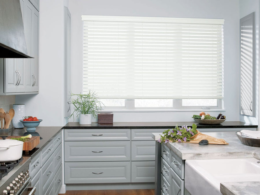 Modern kitchen with light gray cabinets, black countertops, and a large window featuring Everwood® Alternative Wood Blinds with PowerView® Motorization. Fresh vegetables, a potted plant, and kitchenware add stylish touches to the counters.