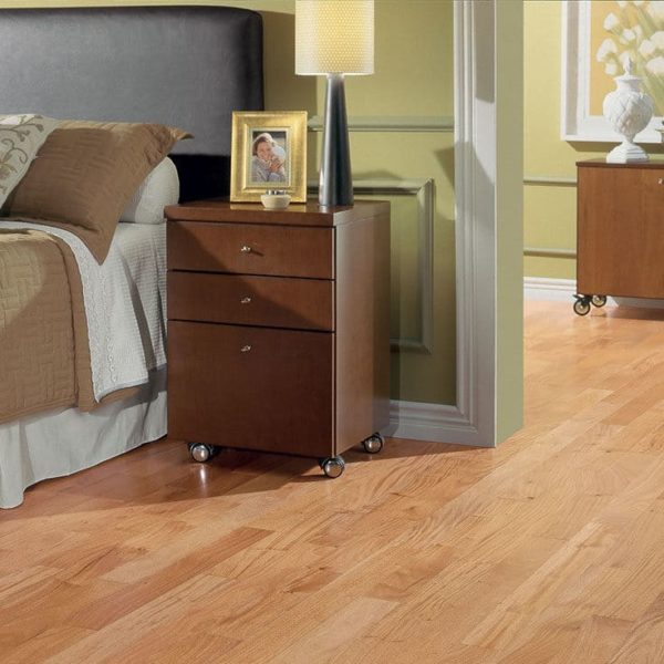 A bedroom featuring Prime Exotic 1/2" x 5 1/4" Hardwood flooring, a brown wheeled nightstand, lamp, framed photo, bed with brown headboard and neutral bedding—sunlight fills the space.