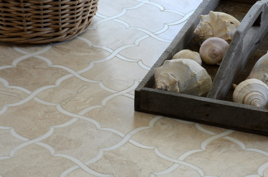 A close-up of Talya Gaia 13 7/16 x 13 7/8 Waterjet Natural Stone tiles featuring modern beige patterns, with a wooden tray of large seashells and part of a wicker basket in the top left corner.
