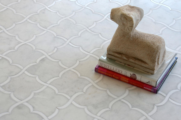 A stone sheep sculpture rests atop two books on a white Talya Gaia 13 7/16 x 13 7/8 Waterjet Natural Stone floor, featuring interlocking curved lines and a blend of marble and modern geometry.
