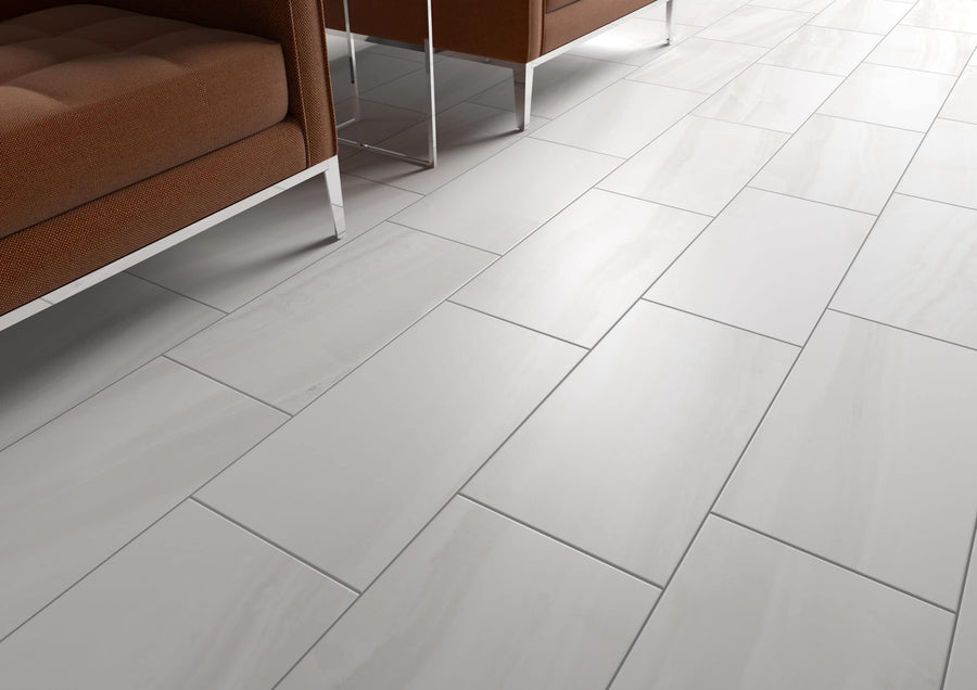 A close-up of Glam 12x24 Matte Porcelain Tile showcases large white rectangles with elegant veining. Brown upholstered chairs with chrome legs appear partially at the image’s edge.