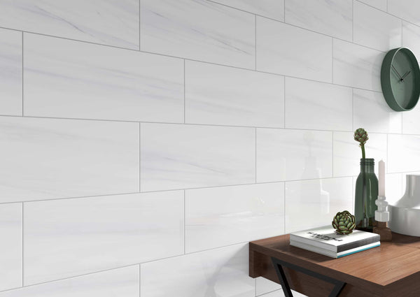 A wall adorned with Glam 4x12 Glossy Porcelain Tiles featuring subtle veining, a green clock, and a wooden table displaying books, green glass bottles, a decorative plant, and white ceramic objects.