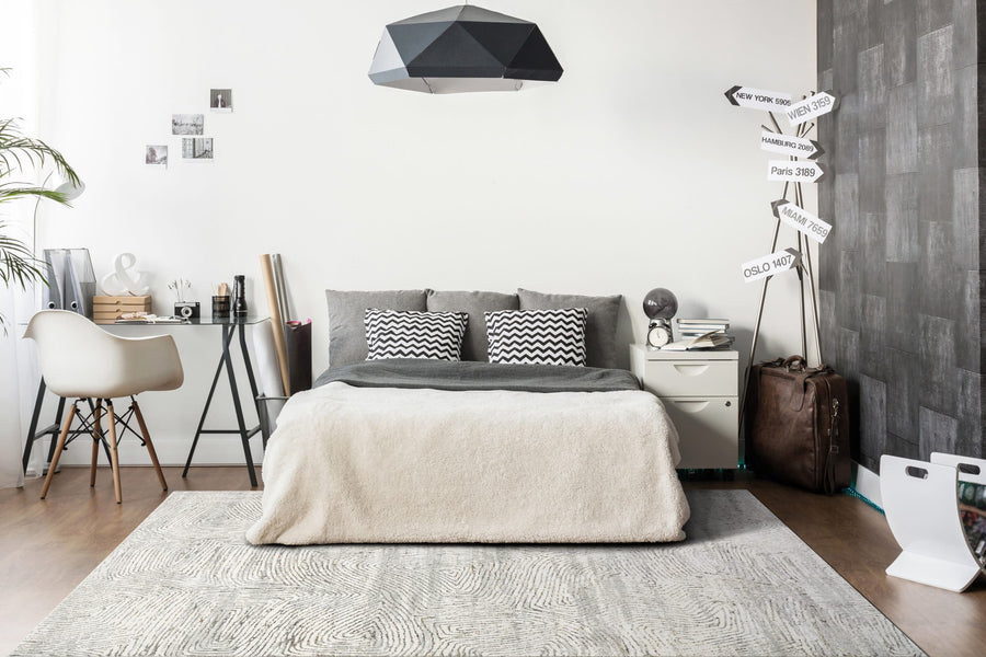 A modern bedroom in gray and white features a bed with patterned pillows, a desk and chair, a geometric ceiling lamp, suitcase, city signpost decor, and the Gold 1351 Area Rug that brings an artistic touch to the space.