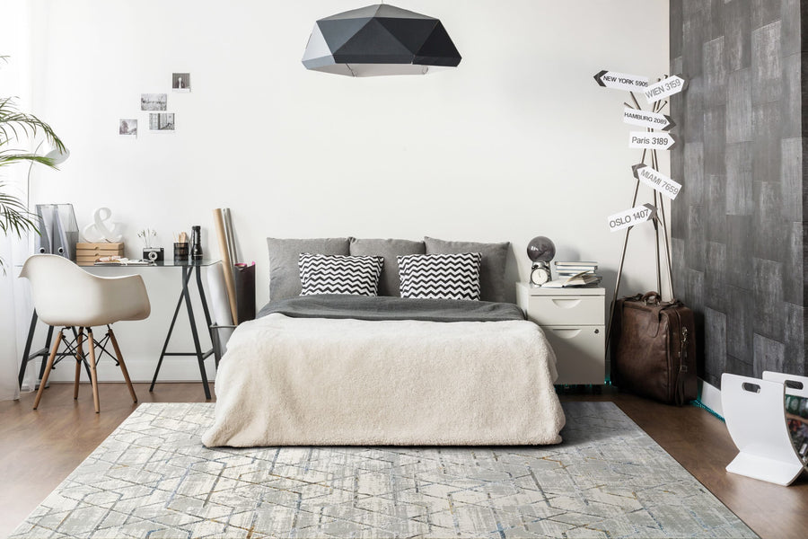 Modern bedroom with a bed, gray and white bedding, geometric lamp, side table, desk, travel signs, suitcase, decorative photos on the walls—all accented by the Gold 1352 Area Rug for a minimalist touch.
