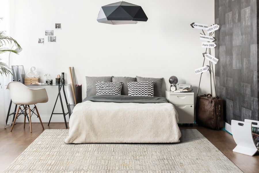 Modern bedroom with a gray bed, chevron pillows, and the Gold 1356 Area Rug. A desk and chair, quirky signpost, soft lighting, and decorative accents—including a suitcase—create a cozy, stylish atmosphere.