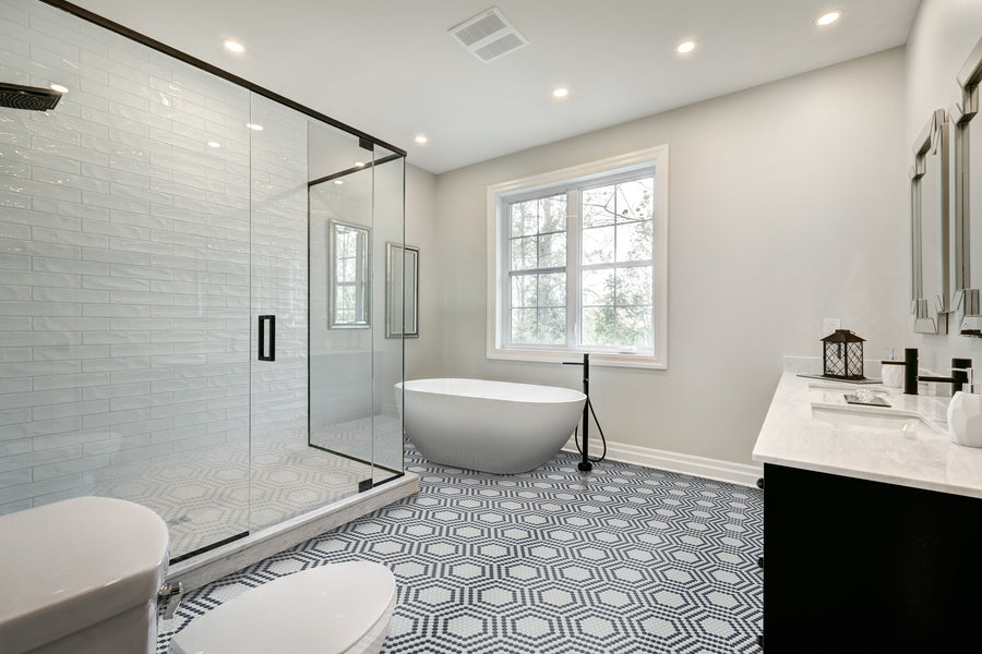Modern bathroom features a spacious glass shower, freestanding white tub, black fixtures, double-sink vanity, and an Evolve Hex Centric Mosaic Tile floor. A large window fills the space with natural light.