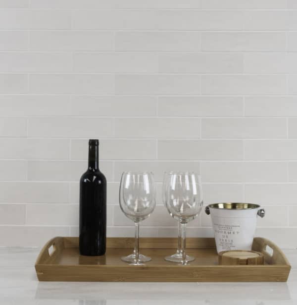 A wooden tray on a white countertop holds a wine bottle, two large glasses, an ice bucket, and a wooden coaster, set against a classic white backsplash tiled with Habitat 2.5x10.3 Subway Tile.