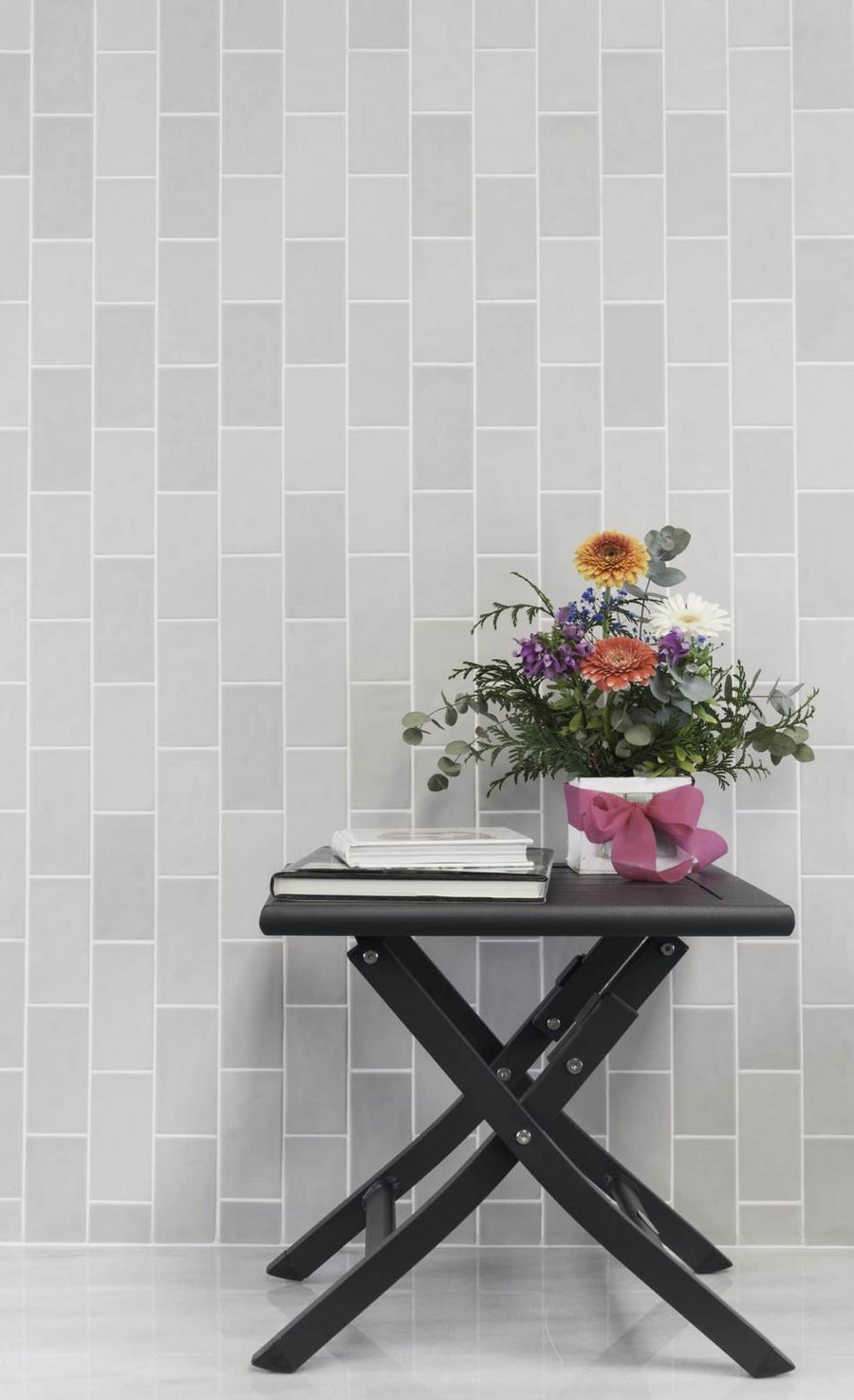 A small black folding table with stacked books and a colorful bouquet in a pink pot stands against a light gray Habitat 2.5x5.1 Subway Tile backsplash.