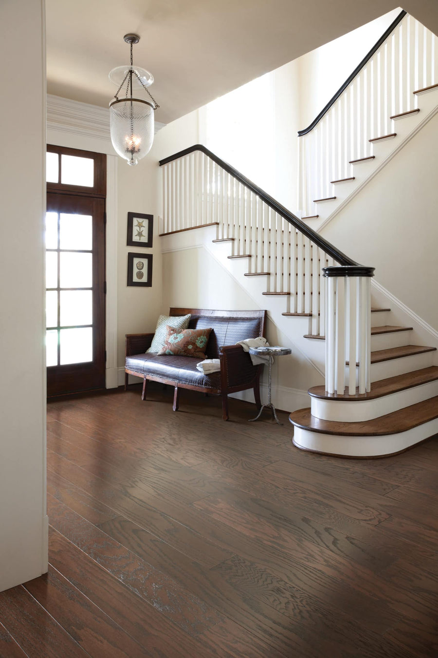 A bright entryway with Homestead 5" Red Oak Hardwood flooring, a cushioned wooden bench, framed wall art, and a curved staircase with white risers and dark handrails. Glass-paneled doors fill the space with natural light.