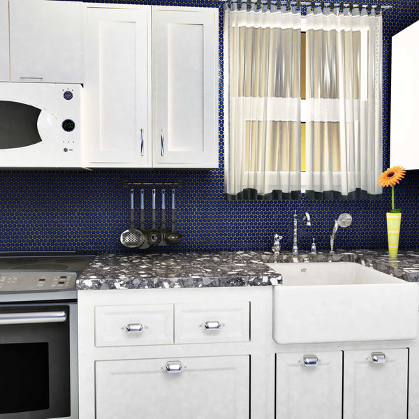 A modern kitchen features white cabinets, a marble countertop, a farmhouse sink, and a stylish Hudson Penny Round Tile backsplash. By the window with sheer curtains are a stove, microwave, knife set, small radio, and an orange flower in a vase.