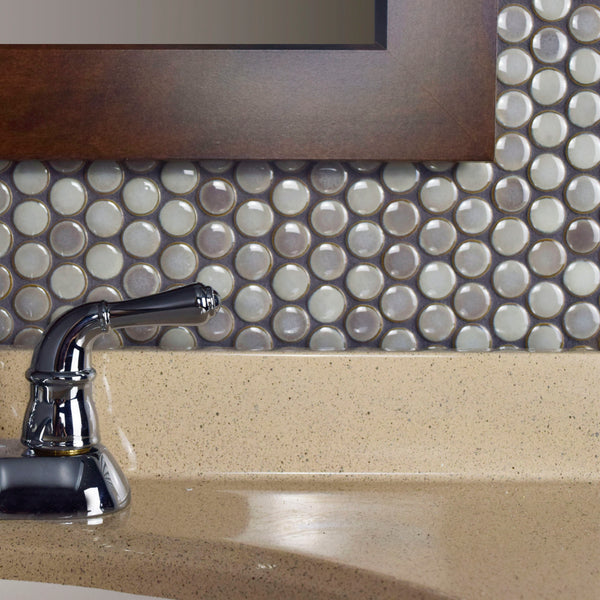 Close-up of a bathroom sink with a chrome faucet, beige countertop, and a wall featuring Hudson Penny Round Tile in glossy gray and white below a wooden-framed mirror.