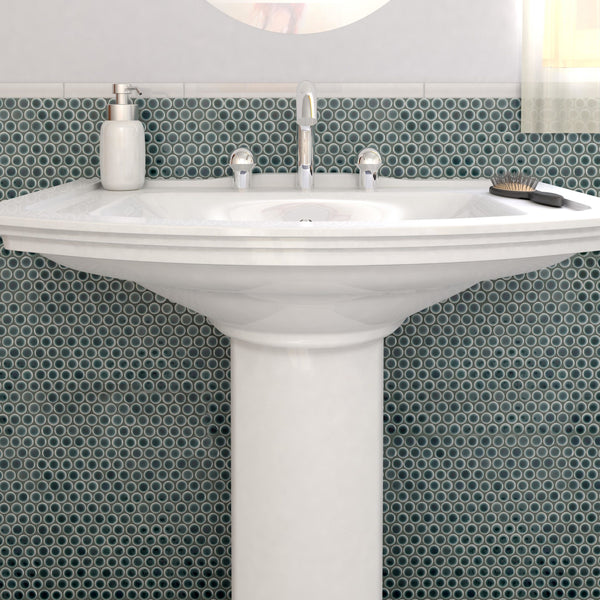 A white pedestal sink with a silver faucet sits before a round backsplash of Hudson Penny Round Tile in teal. A soap dispenser is on the left, a hairbrush is on the right, and sunlight streams in from the window above.