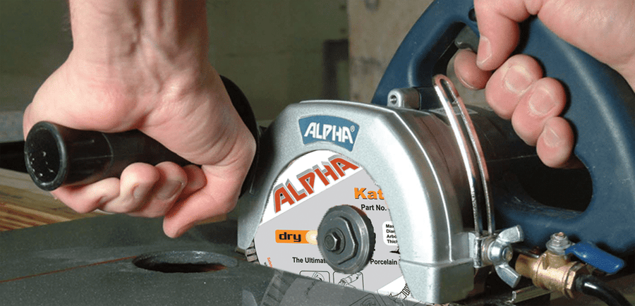 Close-up of hands operating a circular saw equipped with the Alpha Katana Dry Blade for Tile, slicing through material. The gray and blue saw is firmly held—one hand on the handle, the other guiding for precise tile cuts.
