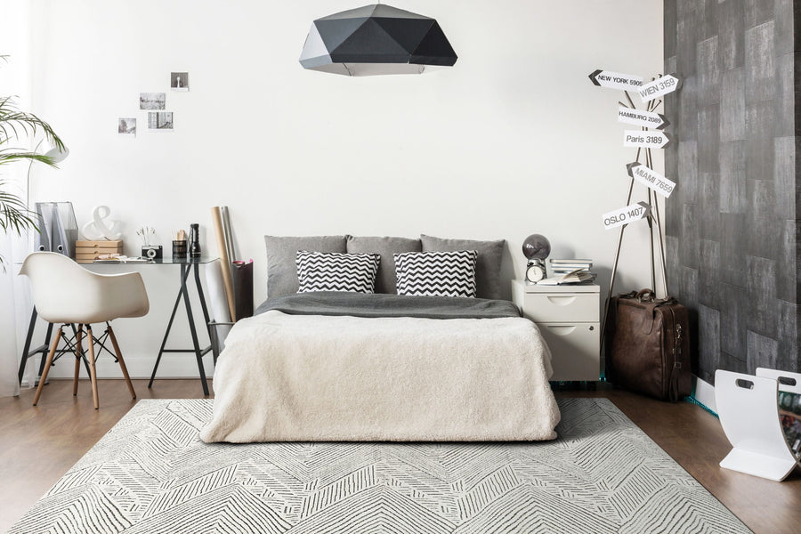 A modern bedroom features a double bed with gray and white bedding, geometric pillows, a small desk and chair on the left, nightstand with travel-themed decor on the right, and the stylish Lotus 8142 Area Rug with high-low texture.