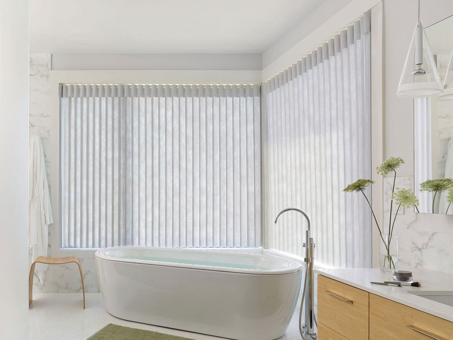 A bright, minimalistic bathroom features a freestanding bathtub, Luminette® Privacy Sheers on corner windows, a light wood vanity with mirror, green rug, and decorative flowers in a vase.