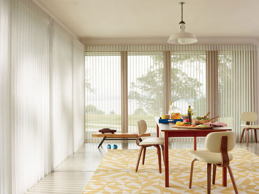 A bright dining room with large windows features Smart Blinds & Shades with PowerView Automation. A wooden table set with food and drinks is surrounded by white chairs on a patterned yellow rug, while trees can be seen outside.